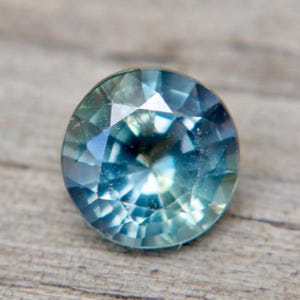 May include: An oval-cut gemstone with a multifaceted surface. The gem displays a range of blue and teal hues, with light reflecting off the cut facets. The gemstone is set against a neutral, wooden background.