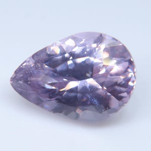 May include: A pear-shaped, faceted gemstone in a light lavender-purple hue. The stone features a brilliant cut with multiple triangular and kite-shaped facets that reflect light, creating a sparkling, translucent appearance with deep violet tones.