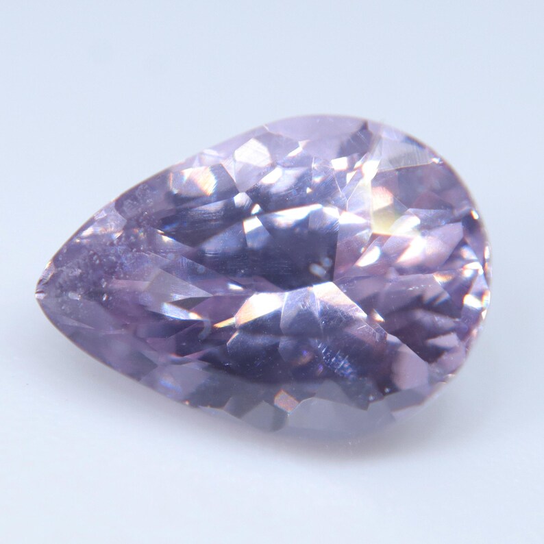 May include: A pear-shaped, faceted gemstone in a light lavender-purple hue. The stone features a brilliant cut with multiple triangular and kite-shaped facets that reflect light, creating a sparkling, translucent appearance with deep violet tones.