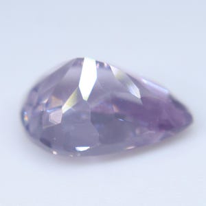 May include: A pear-shaped faceted gemstone with a light lavender purple hue. The stone features multiple triangular and trapezoidal facets that reflect light, creating a polished, translucent appearance with subtle color variations across its surface.