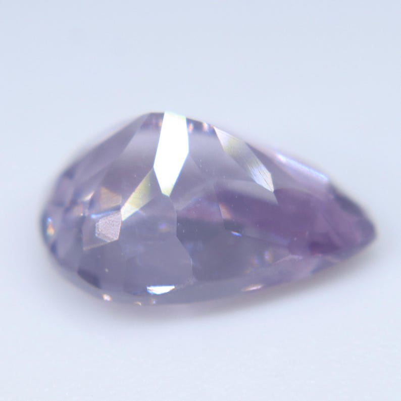 May include: A pear-shaped faceted gemstone with a light lavender purple hue. The stone features multiple triangular and trapezoidal facets that reflect light, creating a polished, translucent appearance with subtle color variations across its surface.