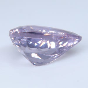 May include: A pear-shaped gemstone with a light lavender purple hue and a faceted surface. The stone features a pointed end and a rounded side, with multiple triangular and diamond-shaped facets that reflect light across its translucent body.