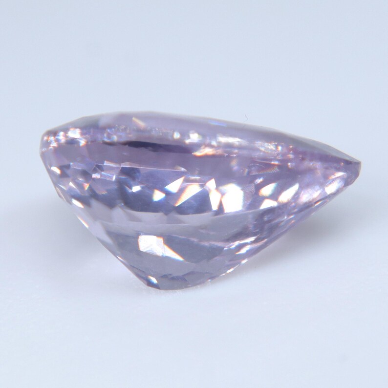 May include: A pear-shaped gemstone with a light lavender purple hue and a faceted surface. The stone features a pointed end and a rounded side, with multiple triangular and diamond-shaped facets that reflect light across its translucent body.