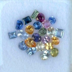 May include: Assortment of loose gemstones in various shapes and colours, including blue, green, yellow, pink, and purple. The gemstones are displayed on a white surface.