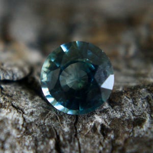May include: A round, faceted gemstone with a deep blue-green color. The gemstone is set against a rough, textured background.