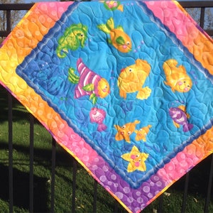 May include: A colorful baby quilt with a blue background featuring a variety of fish, a seahorse, and a starfish. The quilt has a pink, orange, and purple border with white polka dots.