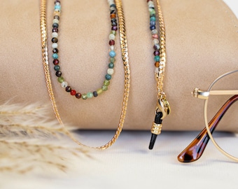 Könnte beinhalten: Eine goldene Kette mit bunten Perlen und einem schwarzen und goldenen Brillenkettenhalter. Die Kette liegt auf einer braunen Oberfläche.