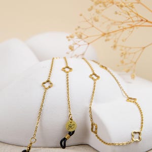 May include: Gold-coloured glasses chain featuring clover-shaped links and a circular charm with the initials "SC". The chain is attached to black glasses holders. The chain is displayed on a white surface.