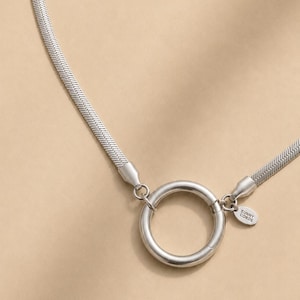 May include: A silver-coloured herringbone chain necklace featuring a central circular ring clasp. A small oval tag attached to the ring is engraved with the text SUNNY CORDS. The chain has a flat, woven texture and connects to the ring via metal end caps.