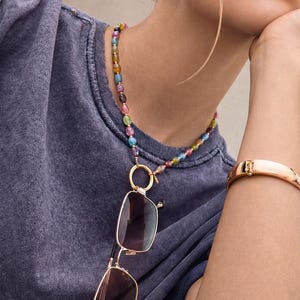 Könnte beinhalten: Eine Perlenkette mit kleinen, mehrfarbigen Edelsteinperlen in Rosa-, Blau-, Grün- und Gelbtönen. Die Kette verfügt über einen goldfarbenen runden Ringanhänger, an dem eine rechteckige Sonnenbrille mit goldfarbenem Rahmen und dunklen Gläsern befestigt ist.