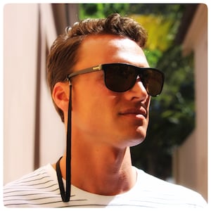 May include: A man wearing black sunglasses with a black strap attached to the frames. The sunglasses have a dark brown lens. The man is wearing a striped shirt.