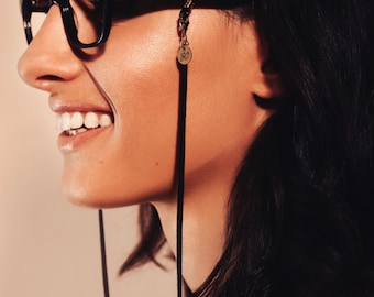 May include: A woman with short black hair wearing black framed glasses with a black leather eyeglass chain. The chain has a small gold charm with the letter "G" on it.