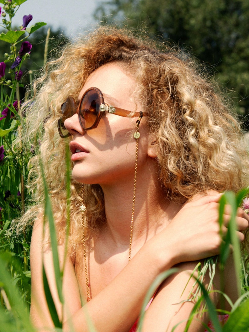 May include: A woman with curly blonde hair wearing brown sunglasses with a gold chain attached to the glasses and a small gold charm hanging from the chain. The woman is wearing a pink tank top and is sitting in a field of green grass.