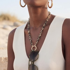 May include: A woman wearing gold hoop earrings and a white ribbed vest top. A necklace with a tortoiseshell chain holds a pair of sunglasses with a green and brown pattern. The sunglasses are hanging from a gold ring.