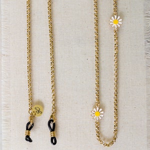 May include: Two gold chains with black rubber ends and small white daisy charms. The chains are on a beige fabric background. One chain has a small gold charm with the letter "G" on it.