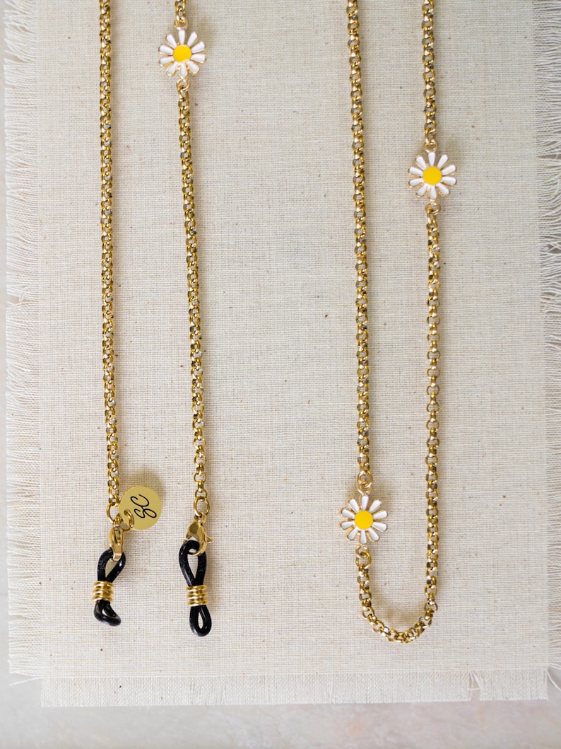 May include: Two gold chains with black rubber ends and small white daisy charms. The chains are on a beige fabric background. One chain has a small gold charm with the letter "G" on it.