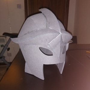 May include: A gray foam helmet with a pointed crown and eye openings. The helmet has a textured surface and a stylized design, possibly for cosplay or costume use. The helmet is sitting on a brown surface.