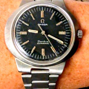 May include: A vintage Omega Genève Dynamic wristwatch with a black dial and silver-tone hands and markers. The watch has a stainless steel case and bracelet. The dial features the Omega logo and the words "Genève Dynamic".