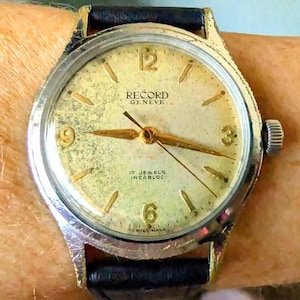 Op de afbeelding: Vintage Record Geneve polshorloge met een goudkleurige wijzerplaat, met de tekst "RECORD GENEVE" en "17 JEWELS INCABLOC". Het horloge heeft gouden wijzers en markeringen, een zilverkleurige kast en een zwarte leren band.