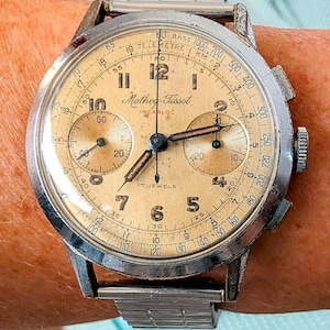 Pode incluir: Relógio cronógrafo Mathey-Tissot vintage com caixa prateada e mostrador creme. O relógio apresenta vários submostradores, uma escala de telêmetro e o texto "17 JEWELS". Pulseira de metal prateado.