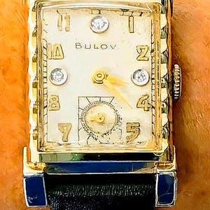May include: A vintage Bulova wristwatch with a rectangular gold-tone case and a cream-colored face. The watch features gold numerals, diamond accents, and a small seconds dial. The black leather band is attached to the gold case with blue accents.