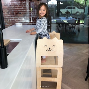 May include: A natural wood-colored toddler step stool with a cat face design. The step stool has a rectangular shape with cut-out sections and a platform for standing. A child in a striped shirt is standing on the step stool near a kitchen counter.