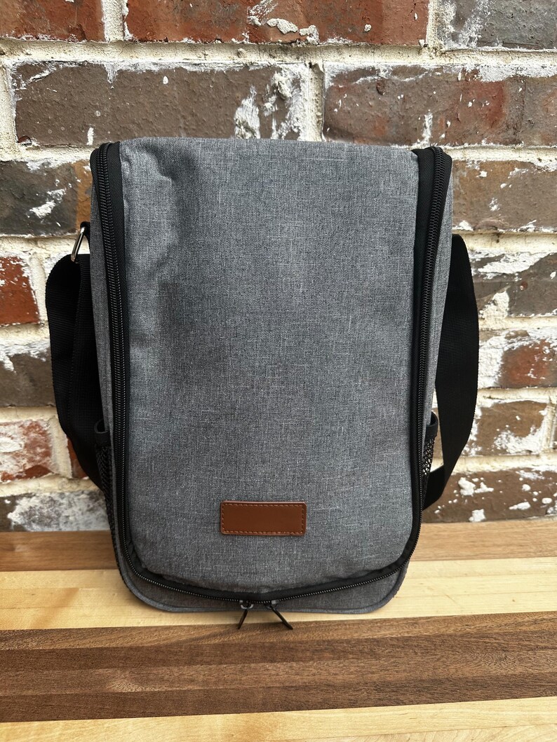 May include: A gray insulated wine tote bag with a brown leather patch and a black zipper. The bag has a black adjustable shoulder strap and a mesh side pocket.