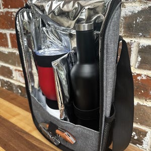 May include: A gray insulated cooler bag with a black shoulder strap. The bag is open and contains a red insulated tumbler, a black bottle, and a wooden corkscrew.