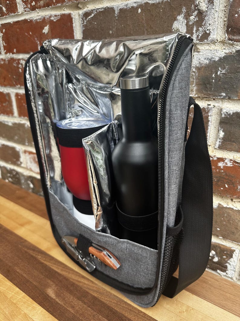 May include: A gray insulated cooler bag with a black shoulder strap. The bag is open and contains a red insulated tumbler, a black bottle, and a wooden corkscrew.
