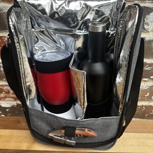 May include: A gray insulated cooler bag with a zipper closure. The bag is open and contains a red insulated tumbler and a black bottle. A silver corkscrew is attached to the bag.