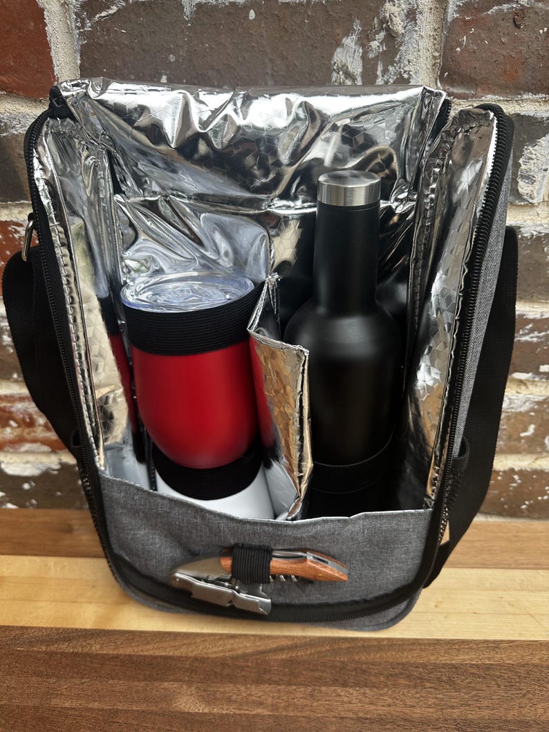 May include: A gray insulated cooler bag with a zipper closure. The bag is open and contains a red insulated tumbler and a black bottle. A silver corkscrew is attached to the bag.
