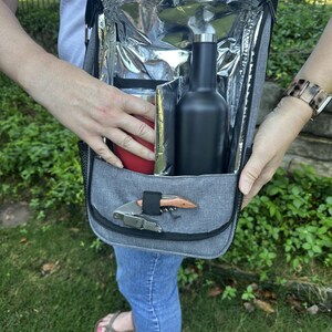 May include: A gray insulated cooler bag with a black zipper and a black bottle inside. The bag has a silver lining and a small pocket on the front with a silver corkscrew.