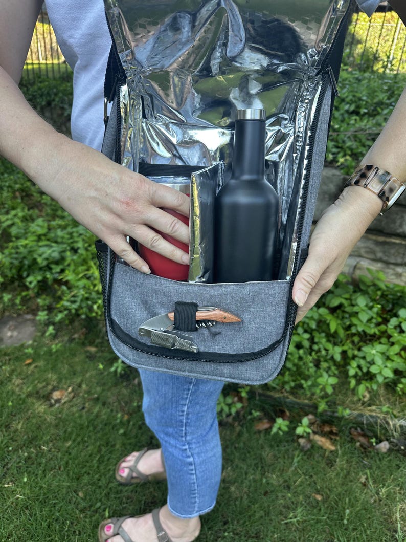 May include: A gray insulated cooler bag with a black zipper and a black bottle inside. The bag has a silver lining and a small pocket on the front with a silver corkscrew.
