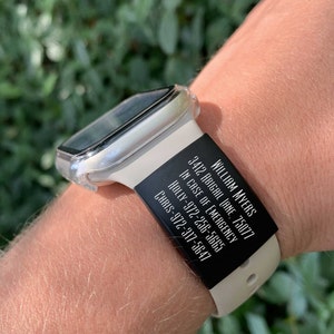 May include: A white Apple Watch with a black silicone band. The band has white text that reads "WILLIAM MYERS", "3412 ABIGAIL LANE, 75077", "IN CASE OF EMERGENCY", "HOLLY-972-256-5665", and "CHRIS-972-317-5647".