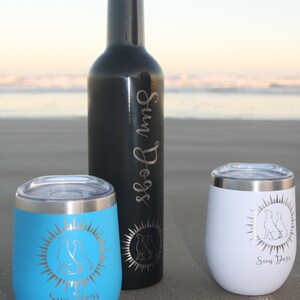 May include: Three stainless steel tumblers with a sun and two dogs design. The tumblers are in blue, white, and black. The black tumbler is a bottle.