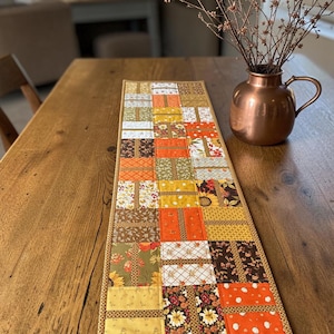 May include: A rectangular quilted table runner featuring a patchwork design of various autumn-themed fabrics in shades of orange, yellow, brown, and green. The fabric patterns include floral prints, small pumpkins, and geometric textures with a border.