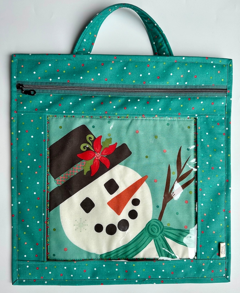 May include: Teal zippered tote bag with a clear window featuring a snowman with a red poinsettia on his hat. The bag has a white polka dot pattern.