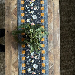 Puede incluir: Un camino de mesa floral blanco y negro con un borde gris y cuadrados naranjas. El camino de mesa est&aacute; sobre una mesa de madera con una planta en una maceta blanca.