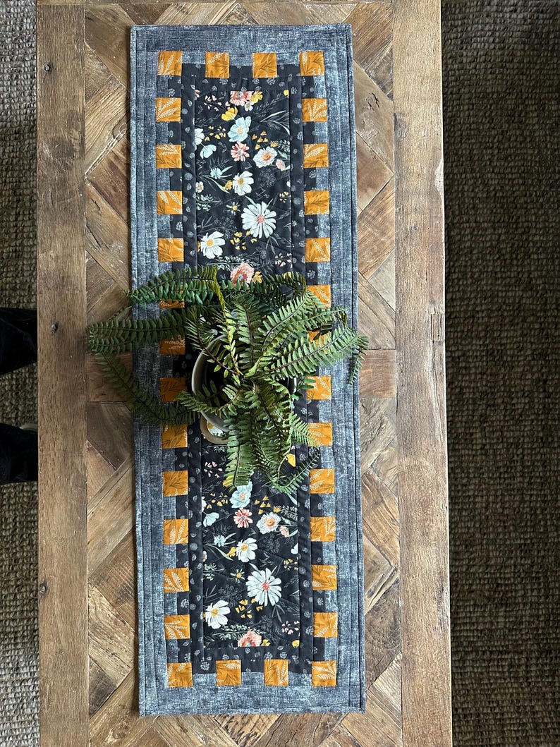 Puede incluir: Un camino de mesa floral blanco y negro con un borde gris y cuadrados naranjas. El camino de mesa est&aacute; sobre una mesa de madera con una planta en una maceta blanca.