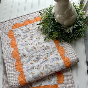 May include: A quilted table runner with a bunny and carrot pattern, featuring an orange scalloped border and a grey gingham edge. A white ceramic bunny statue sits atop a green wreath.