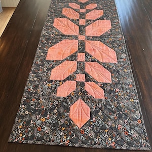Pode incluir: Um caminho de mesa acolchoado retangular com uma coluna central vertical de segmentos de tecido em forma de folha na cor p&ecirc;ssego. O tecido circundante &eacute; cinza-carv&atilde;o escuro com uma estampa floral densa e linhas de acolchoado diagonais.
