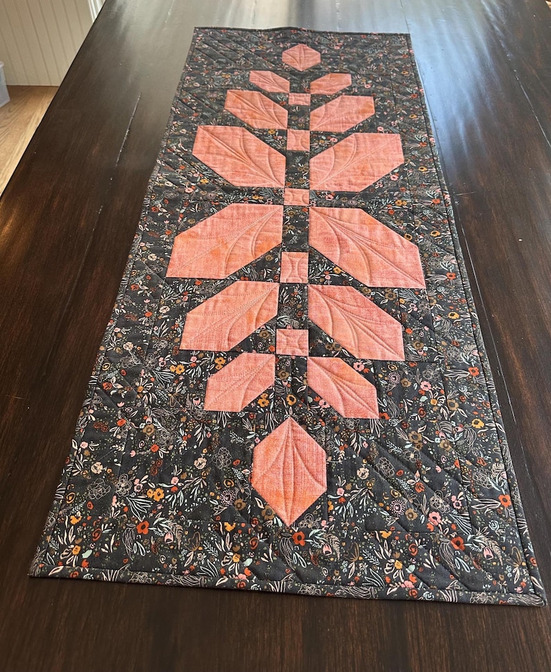 Pode incluir: Um caminho de mesa acolchoado retangular com uma coluna central vertical de segmentos de tecido em forma de folha na cor p&ecirc;ssego. O tecido circundante &eacute; cinza-carv&atilde;o escuro com uma estampa floral densa e linhas de acolchoado diagonais.