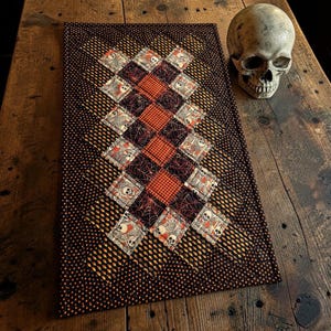 May include: A rectangular quilted table runner featuring a patchwork design of orange and black fabrics. The pattern includes squares with skull motifs, spider webs, and small orange dots on a black background, paired with a realistic human skull.