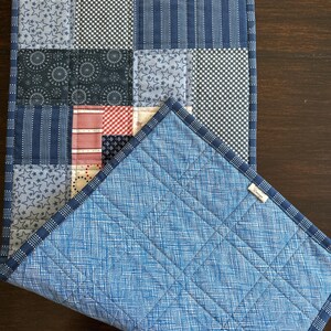 Puede incluir: Camino de mesa patchwork con cuadrados estampados en varios tonos de azul y rojo. El reverso es de tela azul claro con un patr&oacute;n de cuadr&iacute;cula. Los bordes est&aacute;n acabados con un ribete azul oscuro.