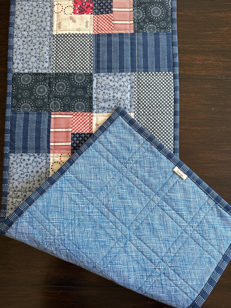 Puede incluir: Camino de mesa patchwork con cuadrados estampados en varios tonos de azul y rojo. El reverso es de tela azul claro con un patr&oacute;n de cuadr&iacute;cula. Los bordes est&aacute;n acabados con un ribete azul oscuro.