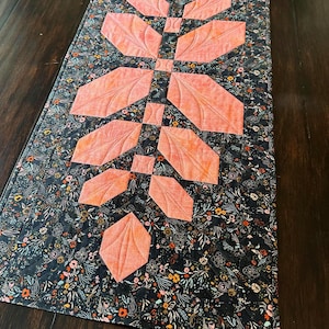 Pode incluir: Um caminho de mesa acolchoado retangular com um fundo de tecido azul-marinho escuro com uma estampa floral densa e multicolorida. Um design central vertical consiste em formas de tecido cor de p&ecirc;ssego dispostas em padr&atilde;o de folha com costura.