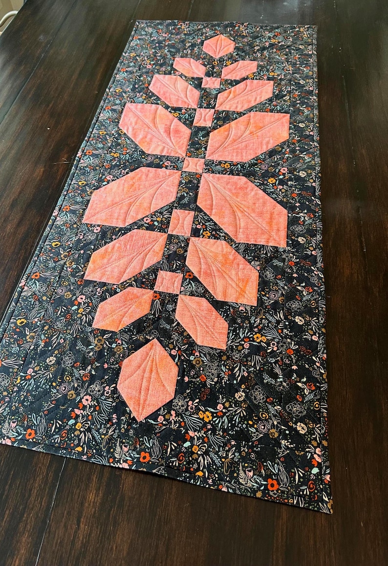 Pode incluir: Um caminho de mesa acolchoado retangular com um fundo de tecido azul-marinho escuro com uma estampa floral densa e multicolorida. Um design central vertical consiste em formas de tecido cor de p&ecirc;ssego dispostas em padr&atilde;o de folha com costura.