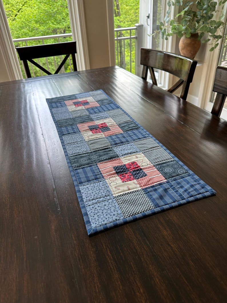Puede incluir: Camino de mesa rectangular de patchwork azul y rojo. El camino de mesa presenta una variedad de telas azules estampadas, incluyendo estrellas, cuadros y rayas, con bloques cuadrados de tela roja y blanca. Se encuentra sobre una mesa de madera marr&oacute;n oscuro.