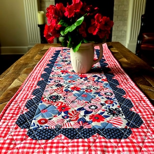 May include: A red and white gingham table runner with a blue and white floral pattern in the centre. The runner has a scalloped edge and is quilted. A white jug with red flowers is on top of the runner.