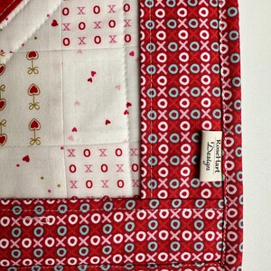 May include: A red, white, and blue fabric quilt with a heart pattern. The quilt is trimmed with a red fabric with a pattern of circles and Xs. The quilt has a label that reads "Rose Hart Design".